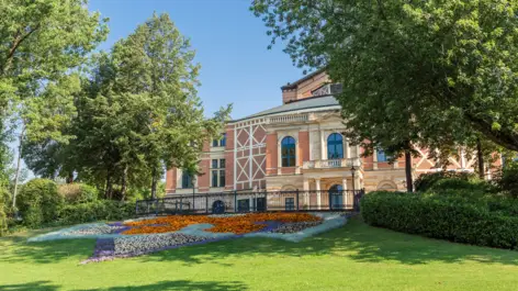 Rote Fassade vom Richard Wagner Haus hinter Bäumen. Davor ein großes, in Muster angelegtes Blumenbeet.