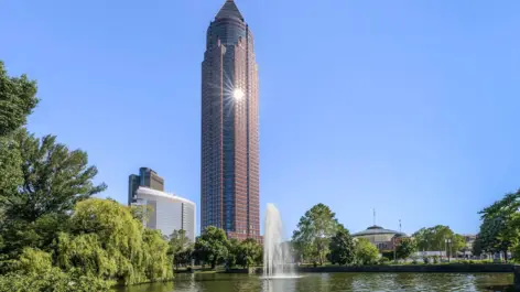 Die Frankfurt Messe wird von der Sonne angestrahlt. Im Vordergrund ist ein großer Teich mit einem Springbrunnen.