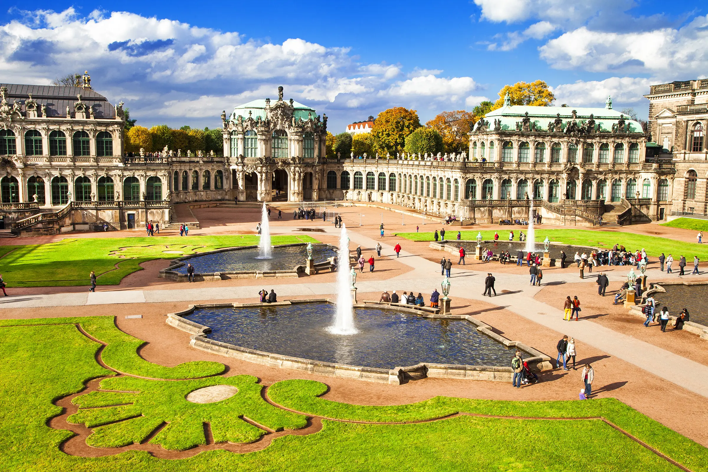 Innenhof vom Zwinger mit zwei Brunnen in denen Fontänen sprudeln- Ringsherum verschiedene, barocke Ornamente als Rasenfläche.