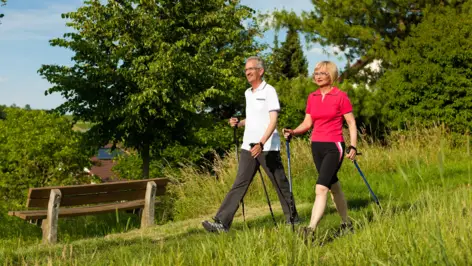 Ein älterer Mann und eine ältere Frau wandern mit Walking Stöcken auf einem Weg.