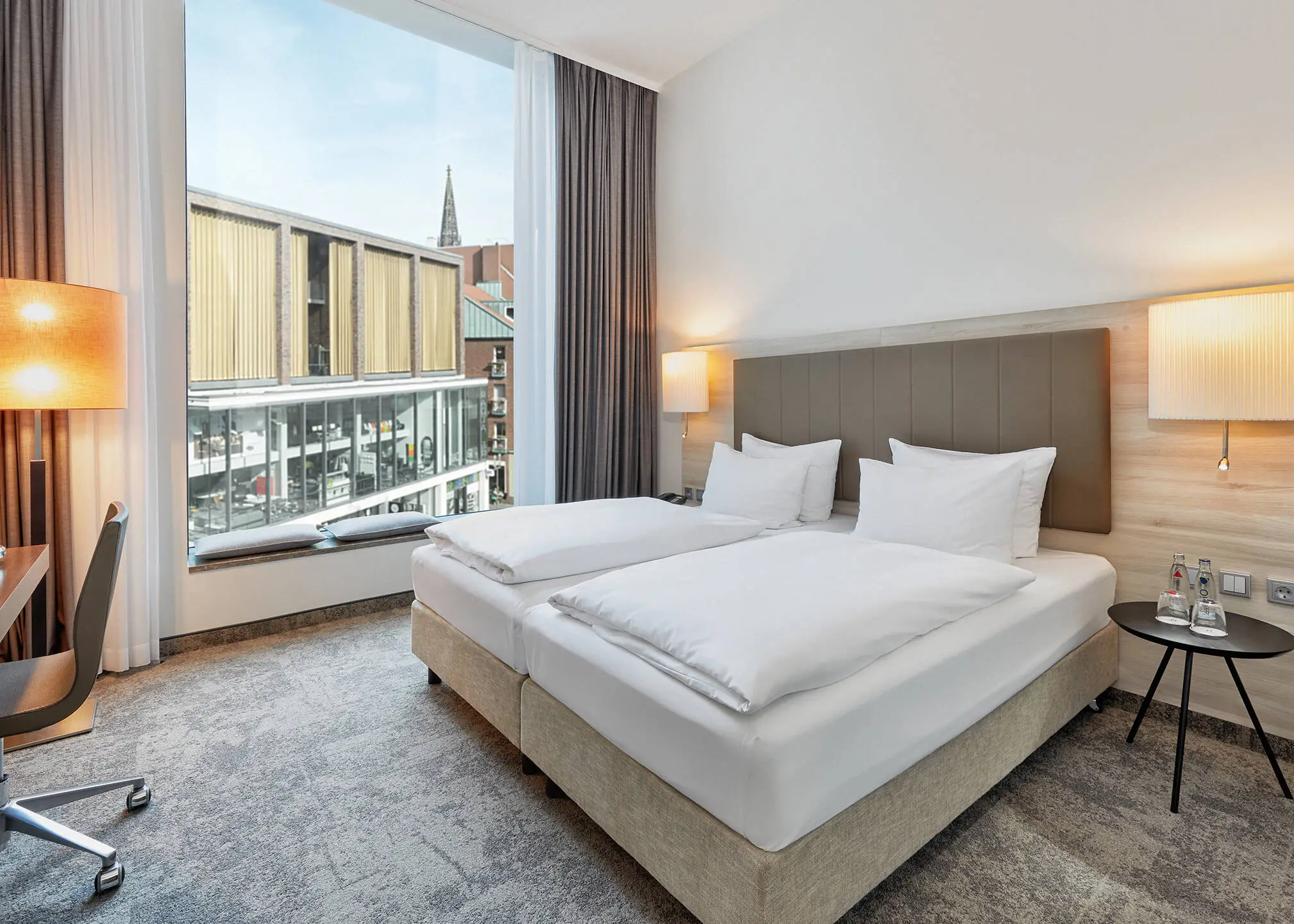 Doppelbett vor einem großen Fenster mit Blick auf die Stadt.