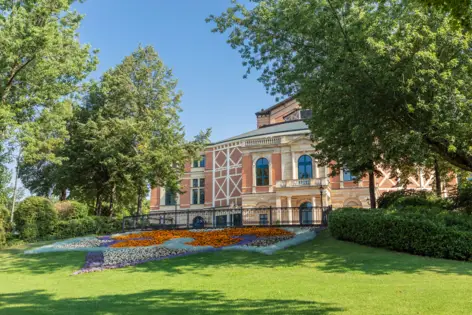 Rote Fassade vom Richard Wagner Haus hinter Bäumen. Davor ein großes, in Muster angelegtes Blumenbeet.