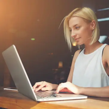 Eine Frau sitzt an einem Holztisch am Laptop und lächelt. Der Raum ist im Hintergrund verdunkelt.