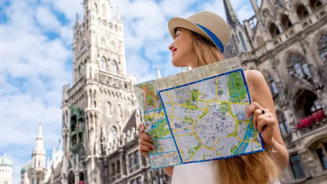 Frau mit Sonnenhut hält einen Stadtplan in der Hand. Sie steht vor dem Münchner Rathaus und schaut es dabei an.