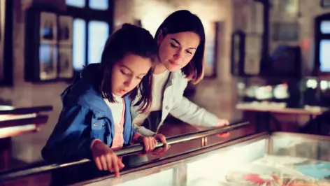 Eine Frau und ein Mädchen stehen vor einer Vitrine in einem Museum und schauen sich etwas an.