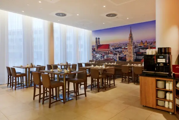 Restaurant mit Sitzplätzen vor großen Fenstern. An der Wand ist eine Fototapete von der Münchener Skyline.