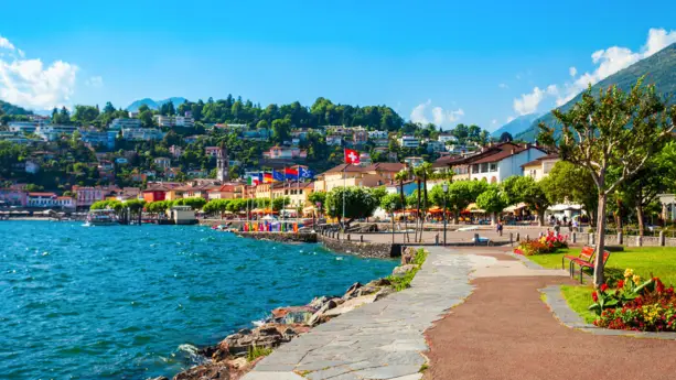 Promenade entlang des Lago Maggiore mit Bäumen und Sitzbänken. Im Hintergrund stehen Häuser und ein kleiner Berg mit Häusern.