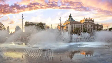 Stachus mit einem großen Wasserspiel während des Sonnenuntergangs. Im Hintergund steht der Justizpalast.