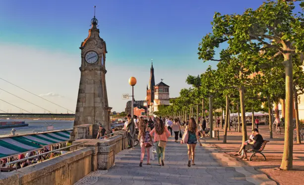 Menschen laufen entlang der Rheinuferpromenade. Im Hintergrund steht der Schlossturm.