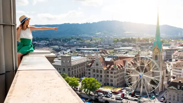 Eine Frau sitzt auf dem Geländer von einem Gebäude und schaut über Zürich.