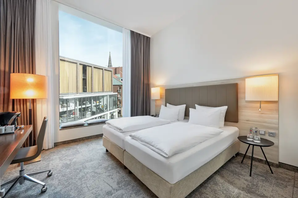 Doppelbett vor einem großen Fenster mit Blick auf die Stadt.