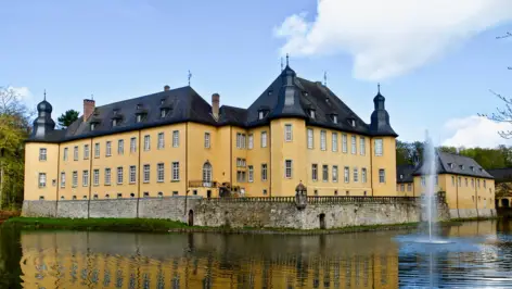 Schloss Dyck umgeben von einem Burggraben gefüllt mit Wasser. Im Wasser ist eine Fontäne.