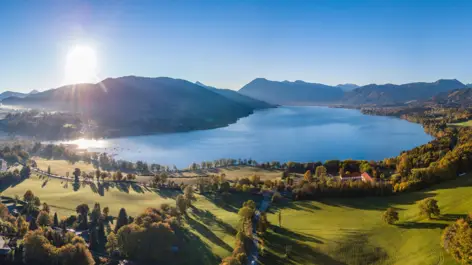 Blick über Wiesen und den Tegernsee. Im Hintergrund geht die Sonne hinter den Bergen unter.