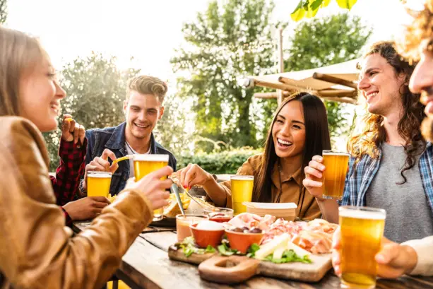 Eine Gruppe von Menschen sitzen lächelnd im Sonnenschein am Tisch und trinken Bier. Auf dem Tisch stehen Bretter mit Aufschnitt.