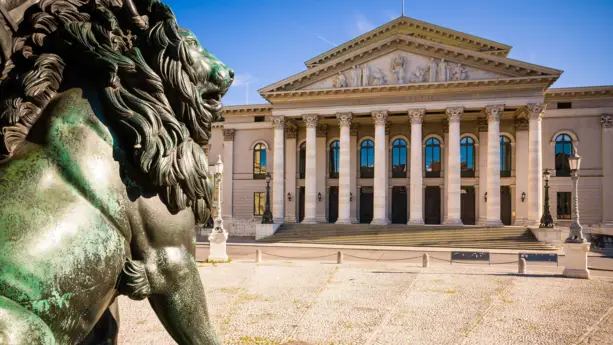 Blick vom Max-Joseph-Platz auf das Bayrisches Nationaltheater. Auf der linken Seite im Vordergrund steht eine Löwenstatue.