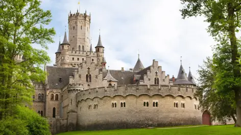 Schloss Marienburg mit seiner Burgmauer. Davor ist eine Wiese mit Bäumen.