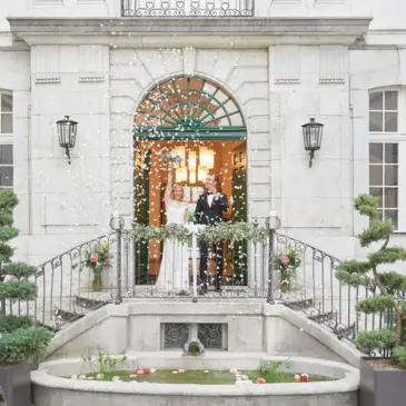 Hochzeitspaar steht vor dem Eingang vom Palais auf eienr kleinen Treppe. Die BRaut hält den Blumenstrauße hoch und es regnen Blüten.