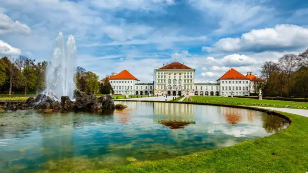 Im Hintergrund steht das Schloss Nymphenburg. Im Vordergrund ist ein großer, runder Brunnen mit einer Fontäne in der Mitte.