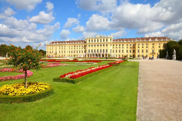 Schloss Schönbrunn im Hintergrund. Vor dem Schloss ist ein großer Schlossgarten mit einer Wiese und verschiedenen Beeten.