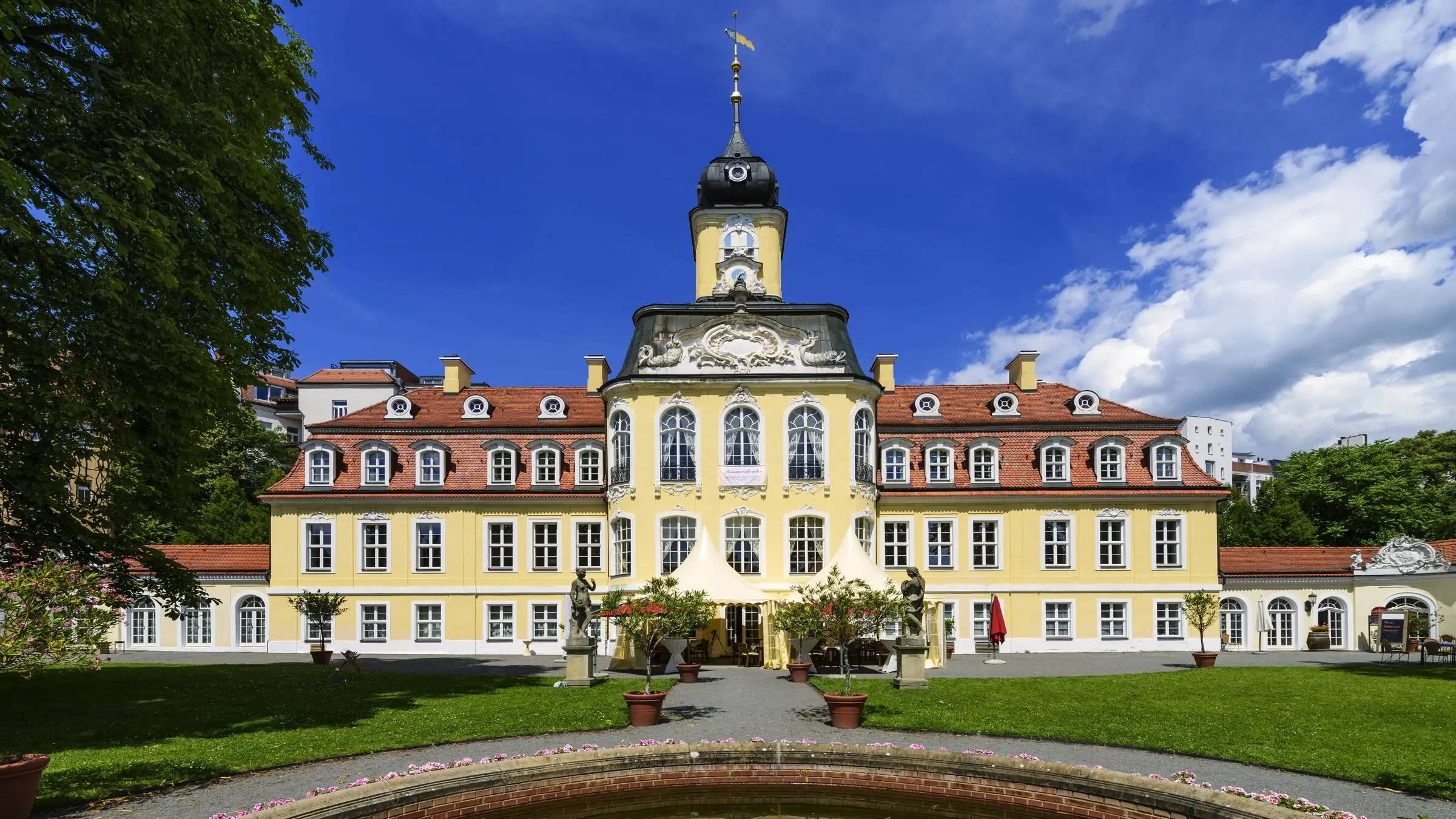 Gohliser Schlösschen mit der gelben Fassade. Davor ein schmaler Weg mit kleinen Bäumen und ein runder Brunnen.