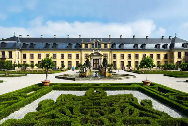 Das gelbe Herrenhaus mit einem Springbrunnen davor. Ringsherum führen Kieswege an kleinen labyrinthähnlichen Hecken.