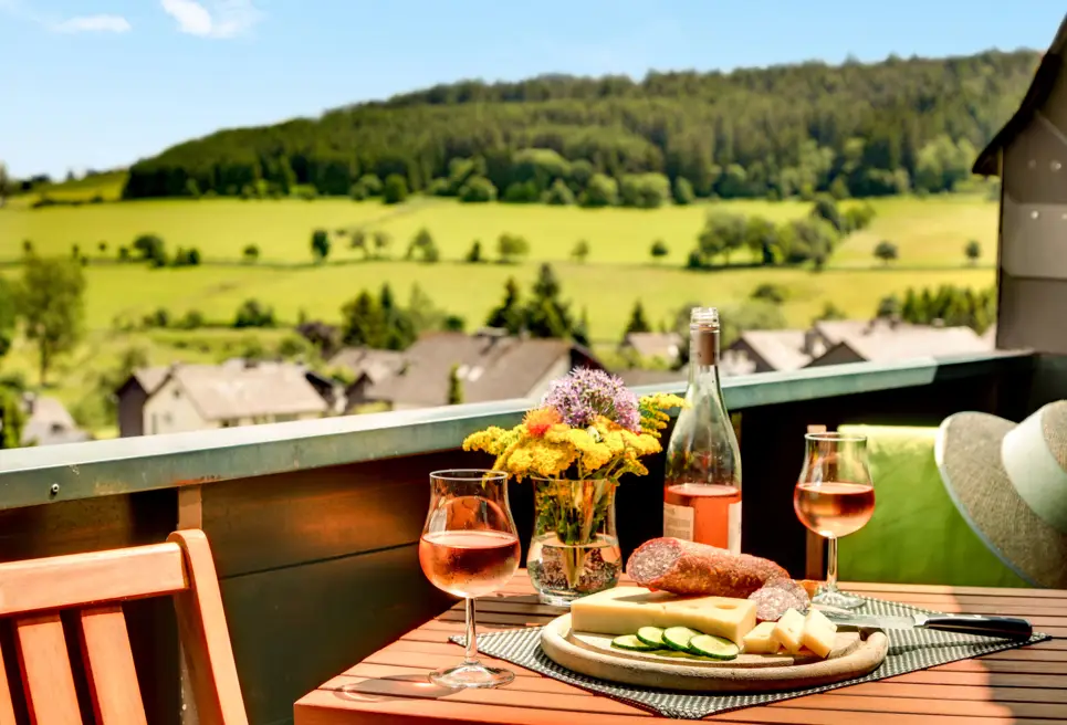 Balkon mit einem kleinen. gedeckten Tisch. Vom Balkon aus sieht man Wiesen und Wälder.