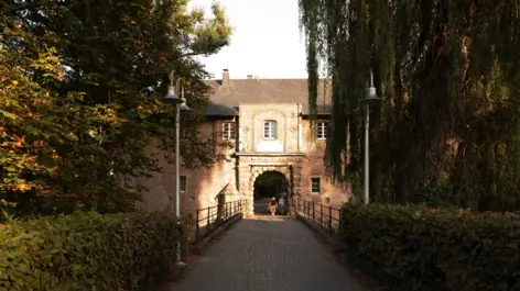 Weg über eine Brück zum Schloss Rheydt. Links und rechts stehen hohe Bäume.