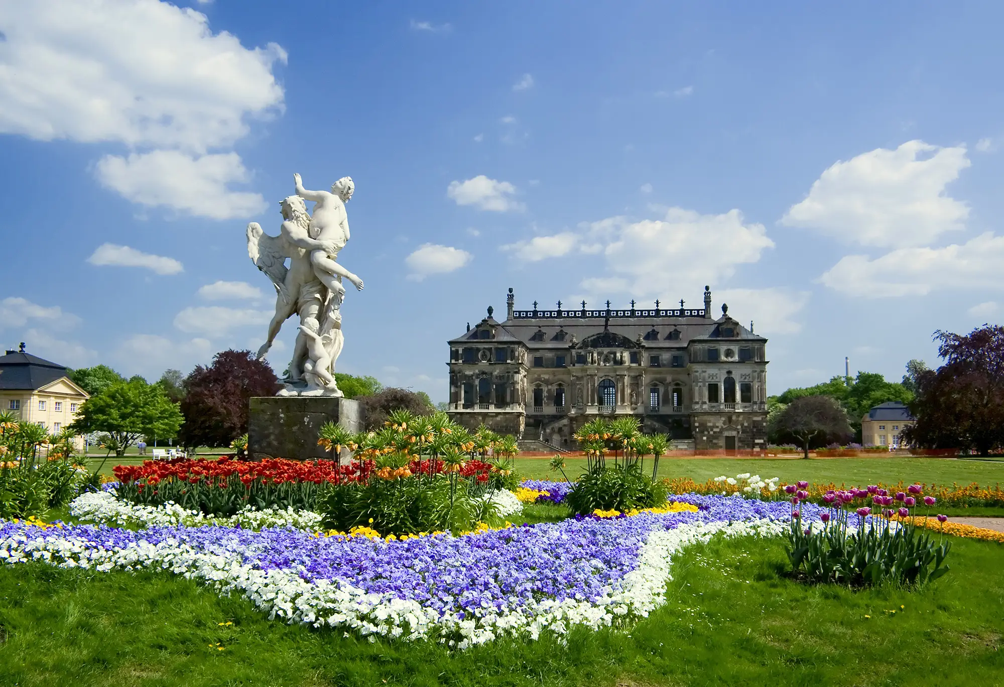 Große Parkanlage mit einem Schloss im Hintergrund. Im Vordergrund ein sternförmiges Blumenbeet mit Engelsstauen in der Mitte.