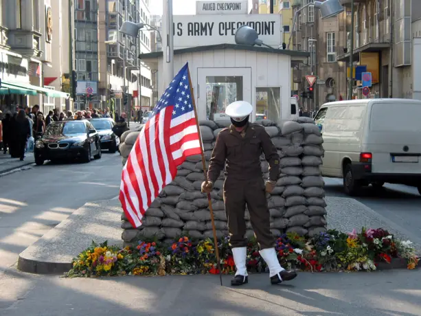 Kleines Wärterhäuschen am Checkpoint Charlie auf der Friedrichstraße. Davor steht ein Soldat und hält eine amerikanische Flagge in der Hand.