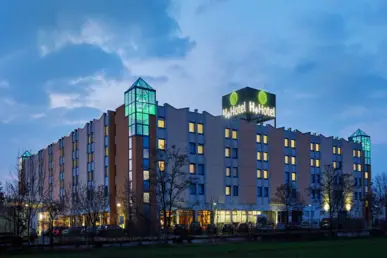 Außenansicht vom Hotel in den Abendstunden.  Auf dem Dach steht ein großer Würfel mit dem Namen H+ Hotel Leipzig-Halle.