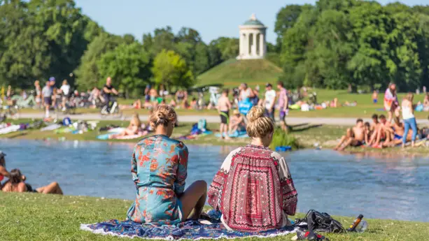 Viele Menschen sitzen an der Isar im Englischen Garten. Im Vordergrund sitzen zwei Frauen, im Hintergrund steht auf einem kleinen Hügel ein Steinpavillon.