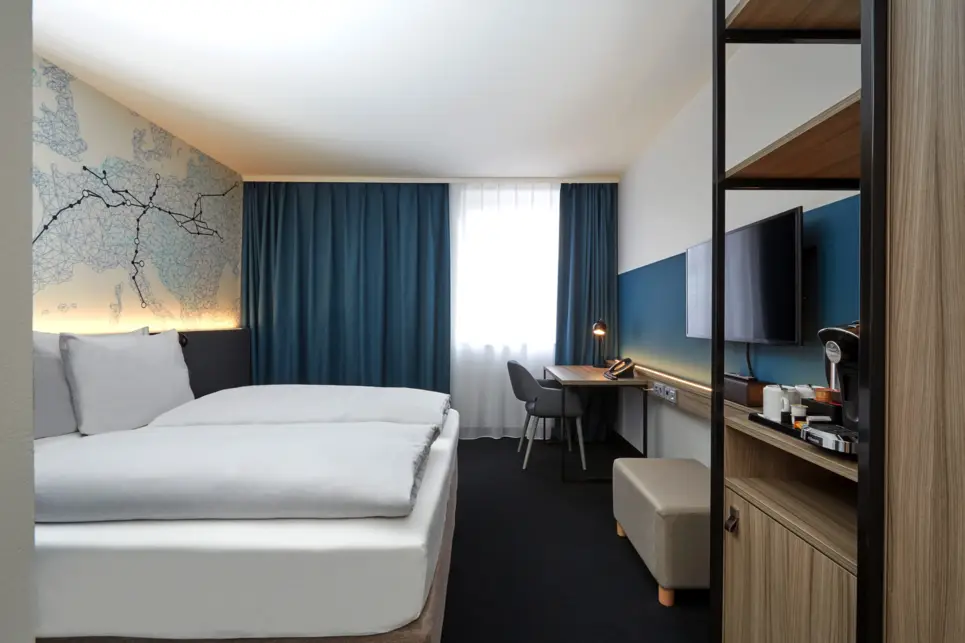 Hotelzimmer mit einem Doppelbett und einem Bild darüber. Auf der rechten Seite ist ein kleiner Schrank und ein Fernseher an der Wand.