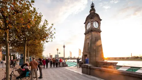 Rheinuferpromenade an der viele Menschen entlang laufen. Im Vordergrund steht eine große Turmuhr und im Hintergrund steht der Rheinturm.