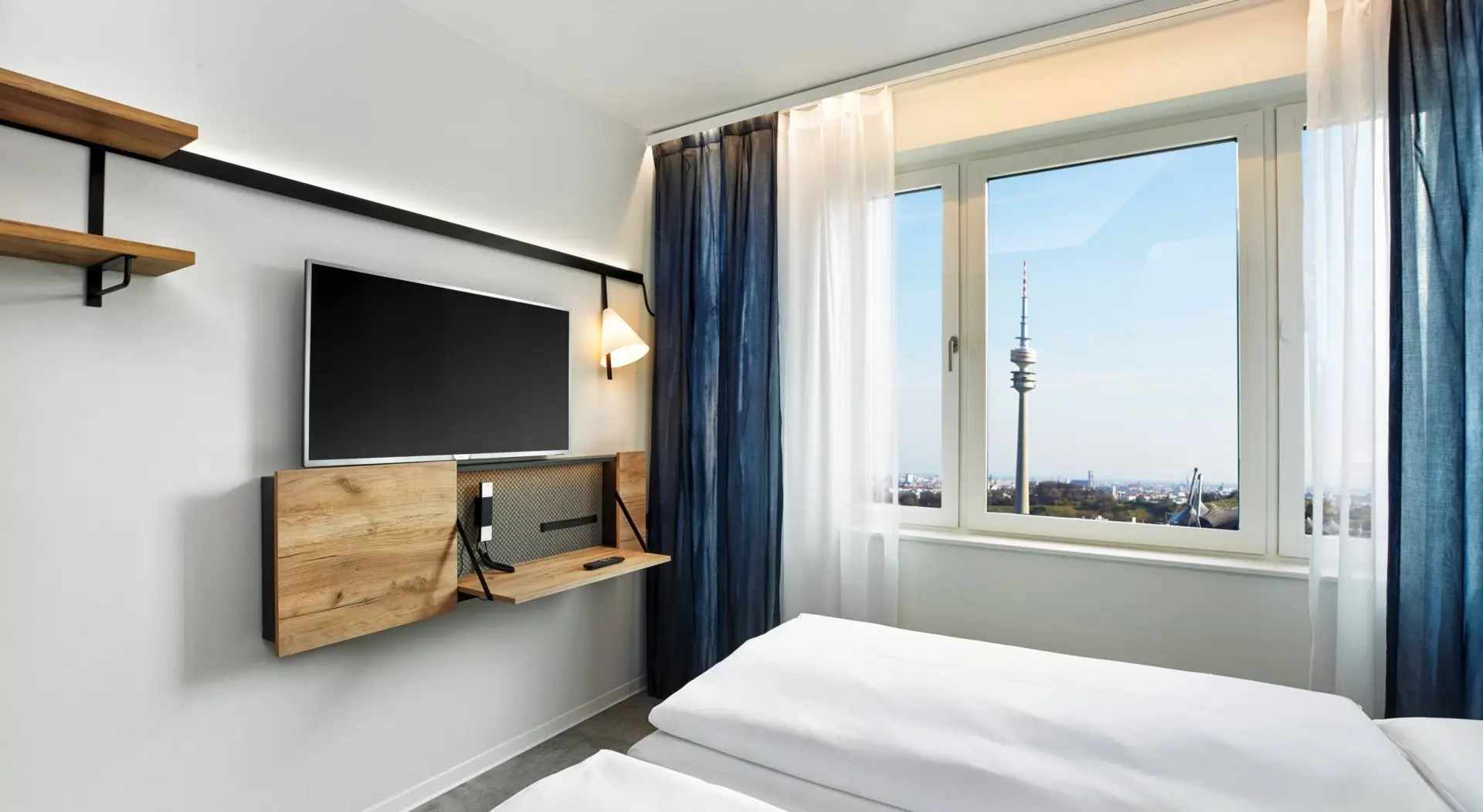 Hotelzimmer mit einem Doppelbett mit Blick auf die Stadt. Vor dem Bett ist ein kleiner Wandschreibtisch und ein Fernseher.