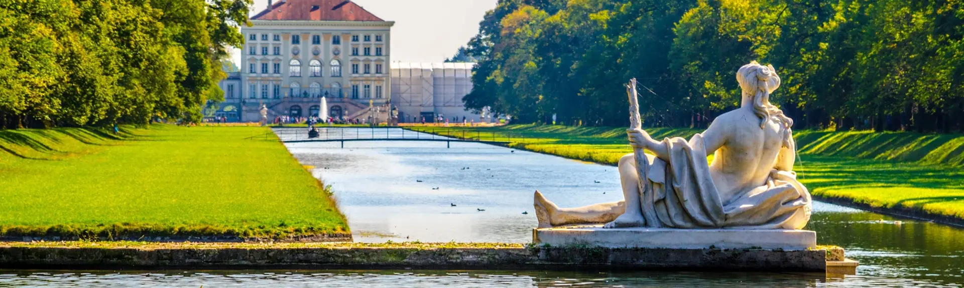 Schlossgartenkanal zum Schloss Nymphenburg. Im Vordergrund liegt eine große Steinnymphenstatue auf einem Steg.