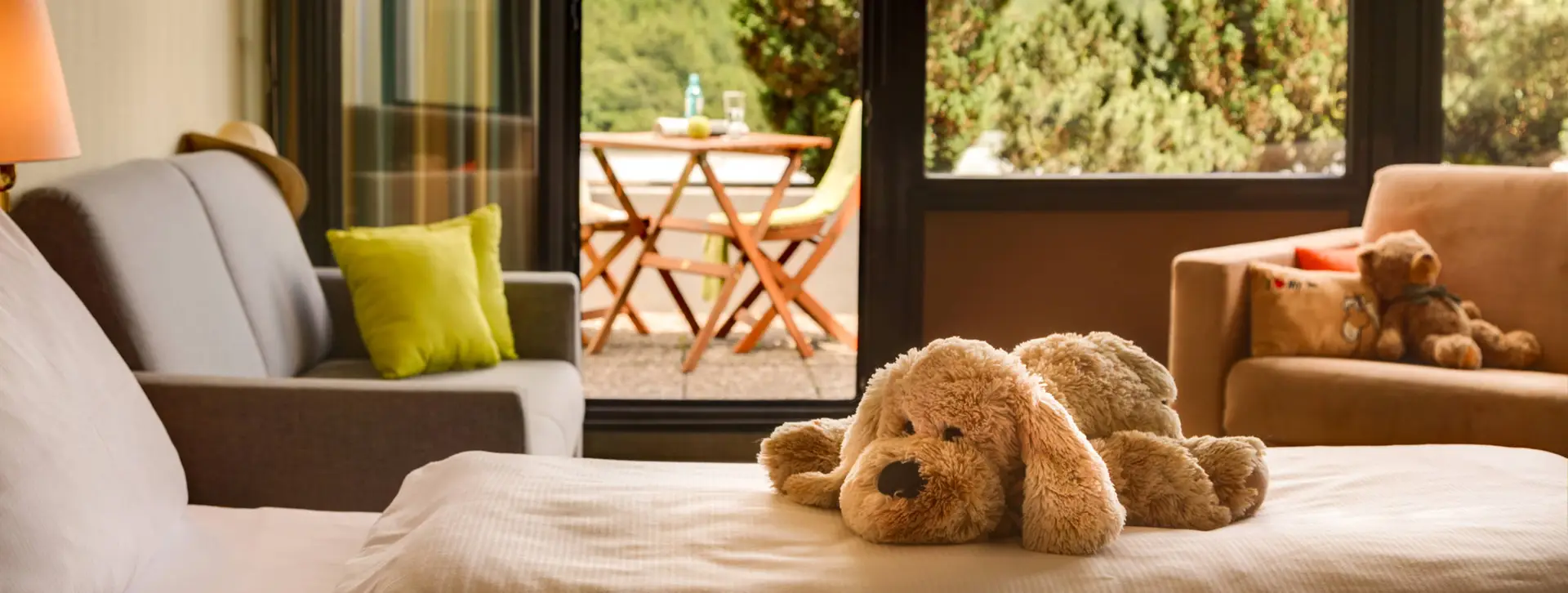 Nahaufnahme auf das Bett mit einem Kuscheltierhund darauf. Im Hintergrund ist der Balkon mit dem Blick auf einen Wald.