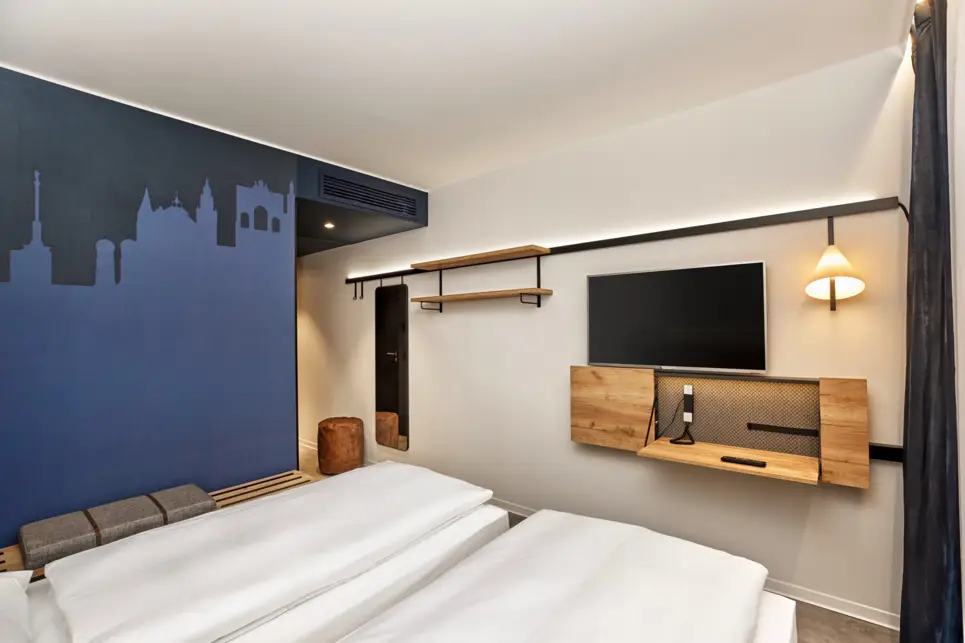 Doppelbett und ein kleiner Schreibtisch mit einem Fernseher im Hotelzimmer. Eine Wand ist in verschiedenen blautönen als Münchener Skyline gestrichen.