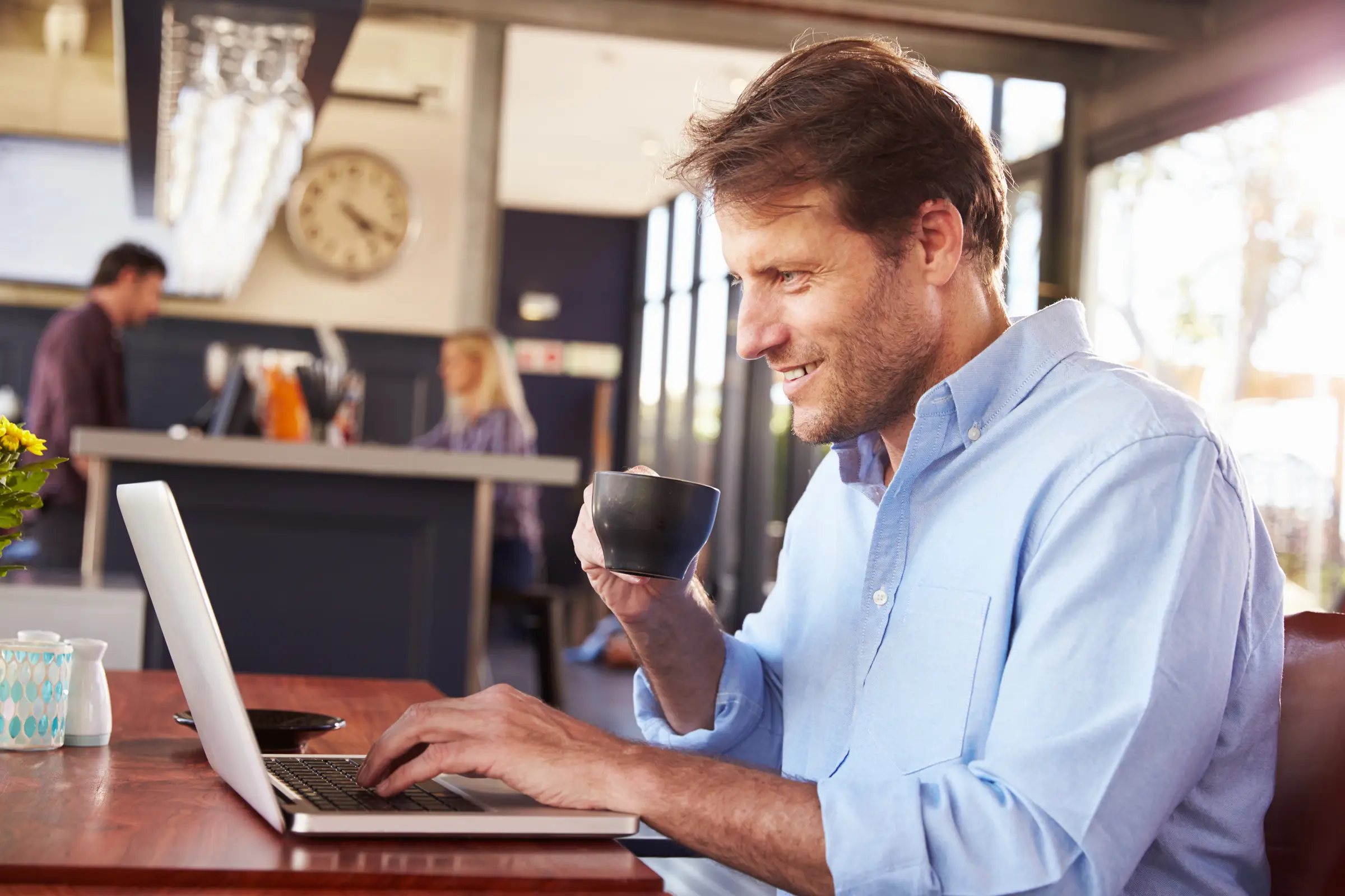 Ein Mann hält eine Tasse in der Hand und schaut lächelnd auf seinen Laptop.