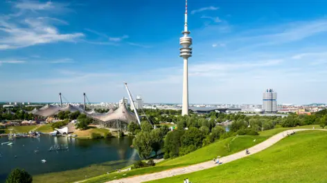 Olympiapark in München mit dem Olympiasee auf der linken Seite und einem Weg auf der rechten Seite. Im Hintergrund steht der Olympiaturm und das Olympiastadion.