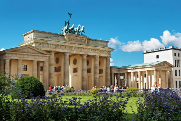 Brandenburger Tor am Tag mit ein paar Menschengruppen davor. Im Vordergrund sind Sträucher und lila Blumen.