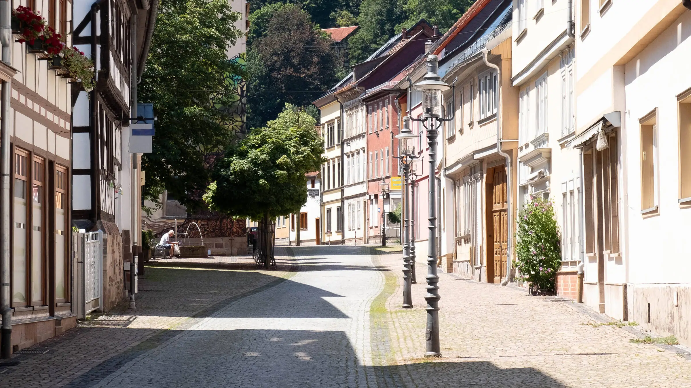 Idílica calle del veraniego casco antiguo de Friedrichroda con casas de entramado de madera y farolillos al borde de la carretera. Al fondo, árboles y un hombre sentado en un banco junto a una fuente. 