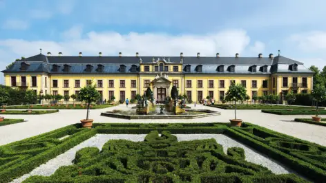 Das gelbe Herrenhaus mit einem Springbrunnen davor. Ringsherum führen Kieswege an kleinen labyrinthähnlichen Hecken.