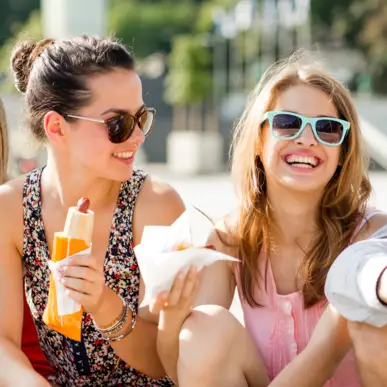 2 Frauen sitzen in der Sonne und lachen. In der Hand halten sie eine Kettwurst.