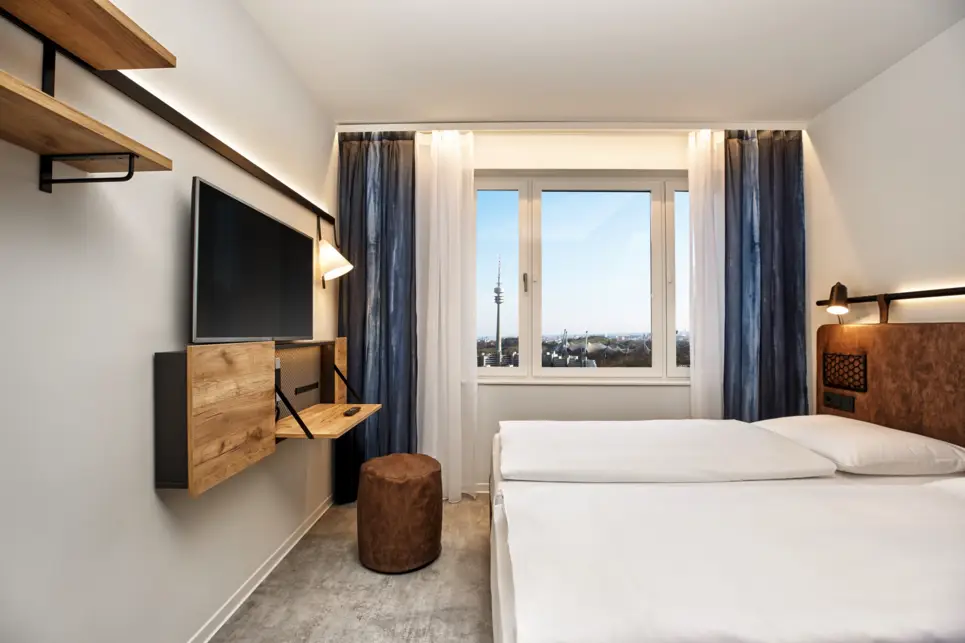 Hotelzimmer mit einem Doppelbett und Blick auf die Stadt. Vor dem Bett ist ein kleiner Schreibtisch und einem Fernseher darüber.