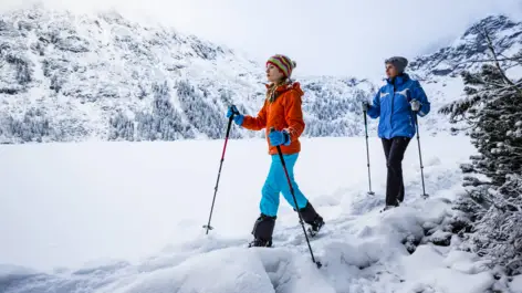 Zwei Skilangläufer laufen durch tiefen Schnee. Im Hintergrund sind Berge.