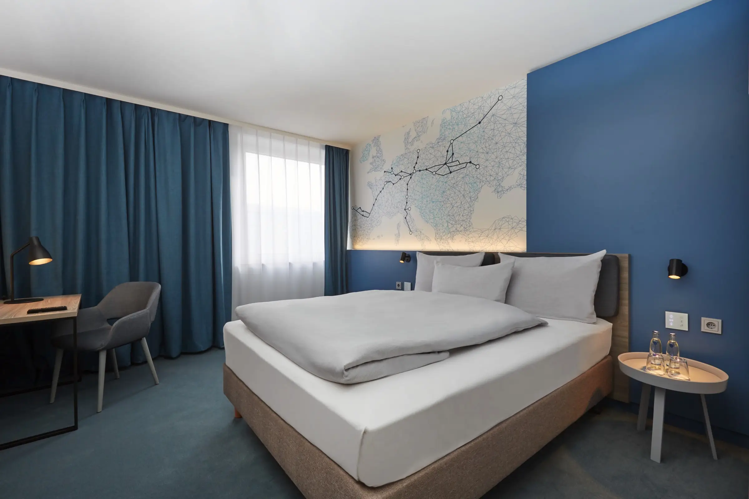 Modern gestaltetes Hotelzimmer mit einer blauen Wand. An der Wand hängt eine in linien gestaltete Europakarte mit dicken Linien abgehend von Leipzig.