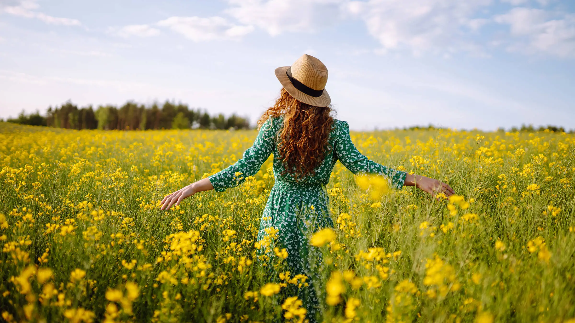 A woman in a straw hat and green dress stands in a rape field.