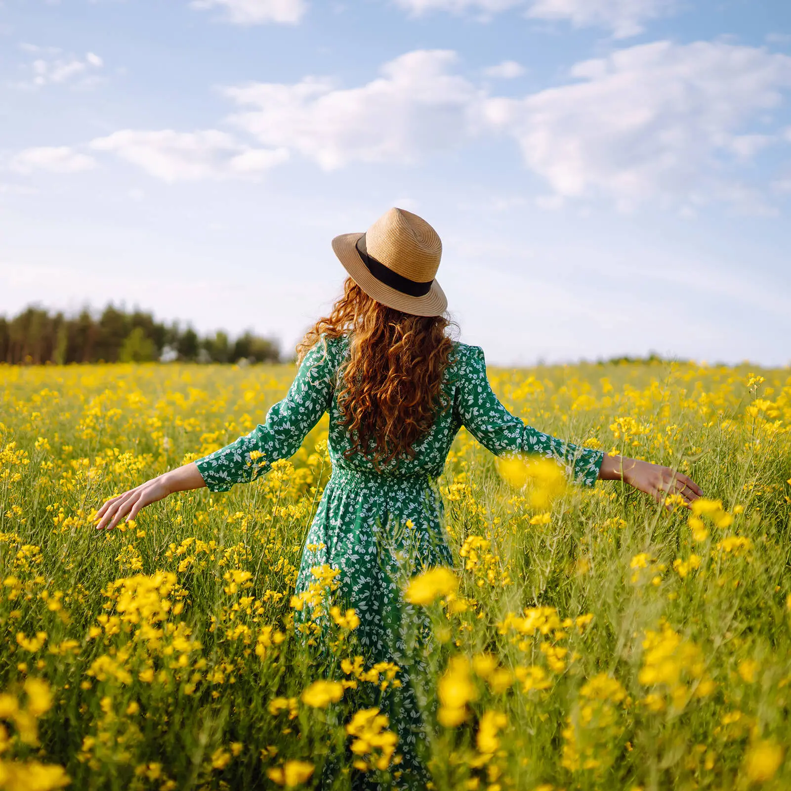 A woman in a straw hat and green dress stands in a rape field.